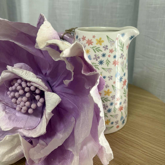 Close-up of a large purple flower with a floral-patterned vase in the background.